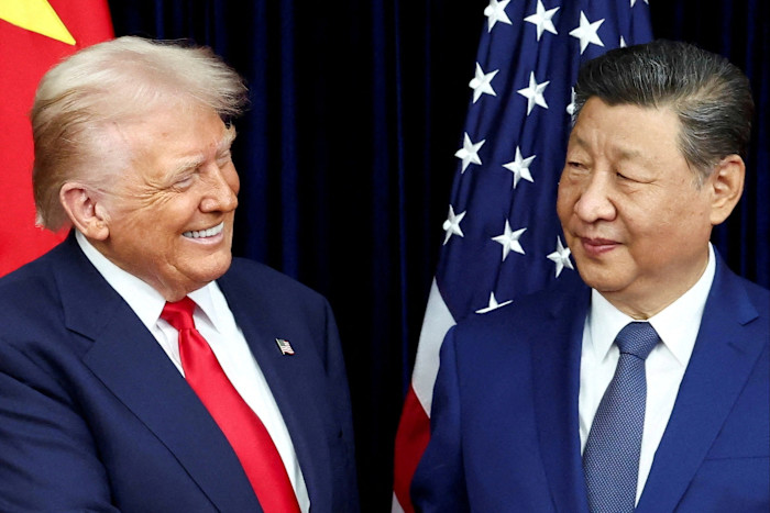 Donald Trump and Xi Jinping face each other and smile during a bilateral meeting, with U.S. and Chinese flags in the background.