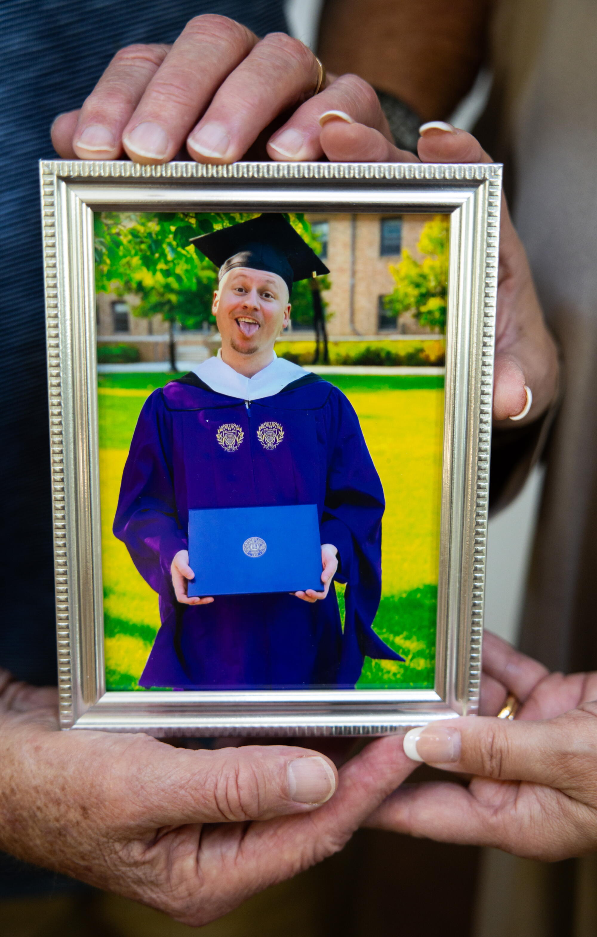 Ryan Regan makes a silly face in a graduation cap and gown, in a framed photo cradled by his parents' hands.