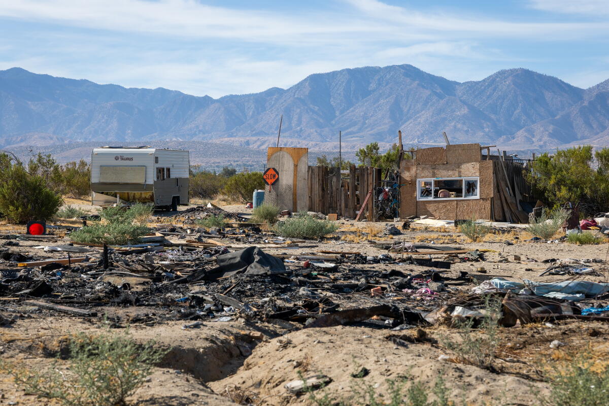 An RV near dilapidated structures with garbage strewn in front and mountains in the background