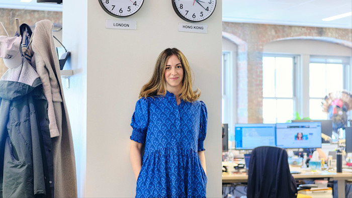 Harriet Clarfelt stands in a blue dress in the FT’s New York office, with clocks showing different time zones on the wall behind her.