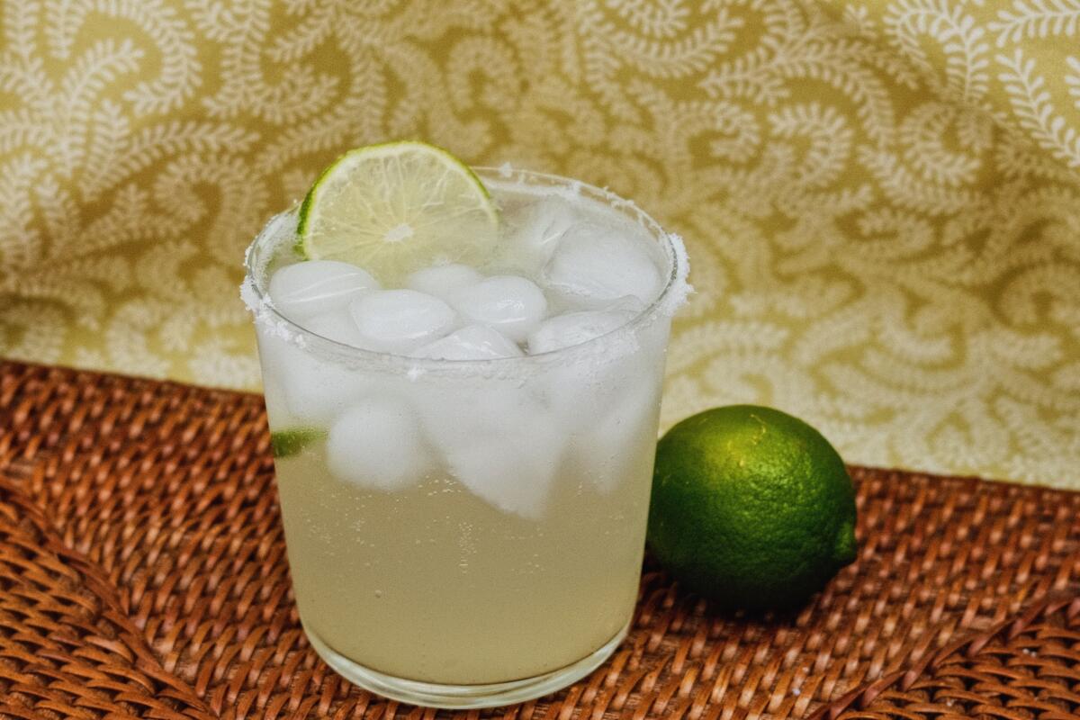 A glass of lime and sparkling water on a brown textured placemat against green patterned fabric. To the right is one lime.