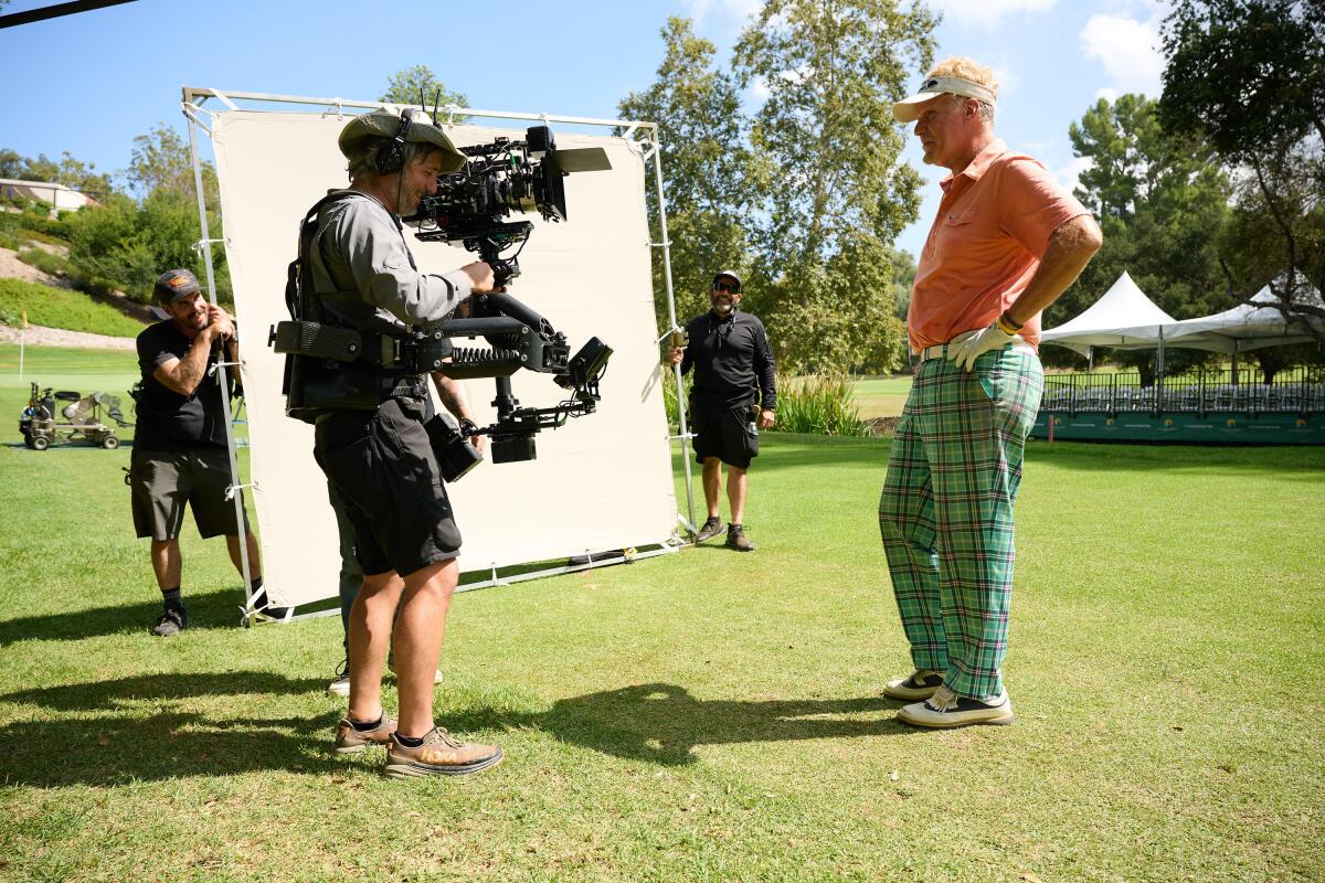 A man holding a movie camera points it toward a man standing on a green lawn.