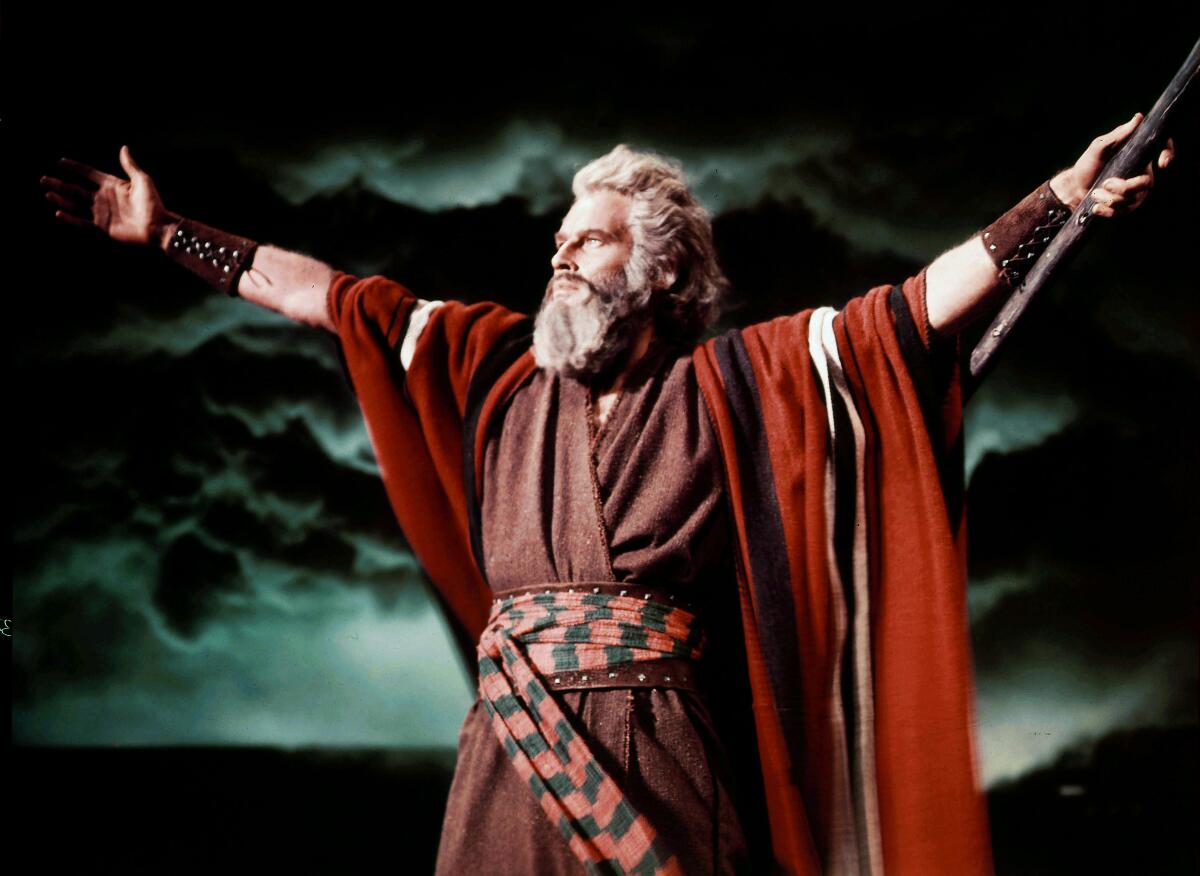 Charlton Heston in robes and a gray beard, holding up both arms. In the background is a stormy sky.