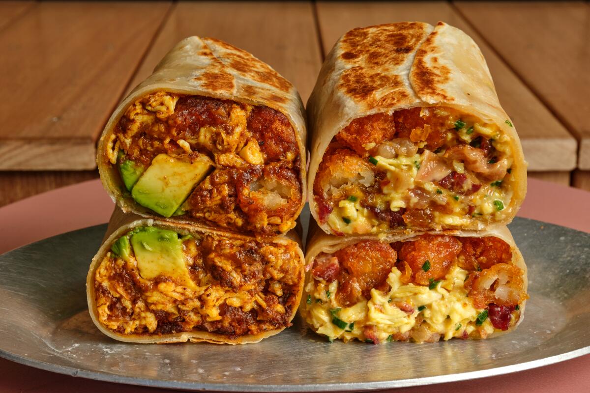 Two burritos with egg, avocado, hash browns and more are cut in half so that their fillings are visible.