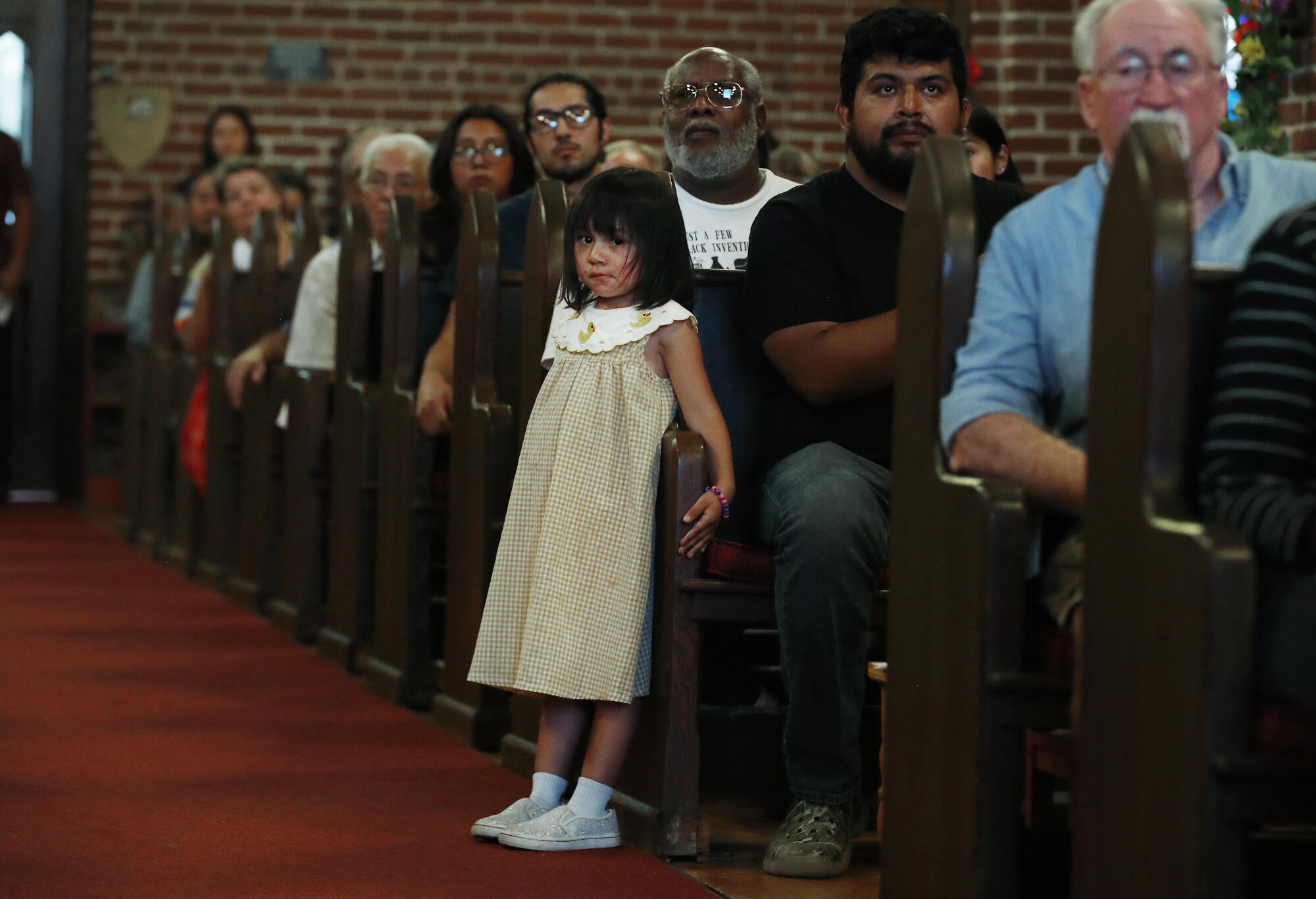 a girl stands next to her father sitting in a church pew with others