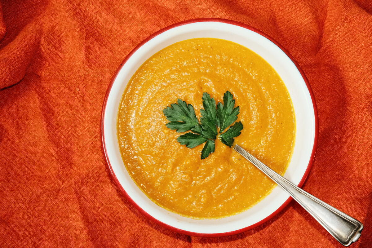 A bowl of vegan carrot and ginger soup in a red-rimmed white bowl on orange cloth.