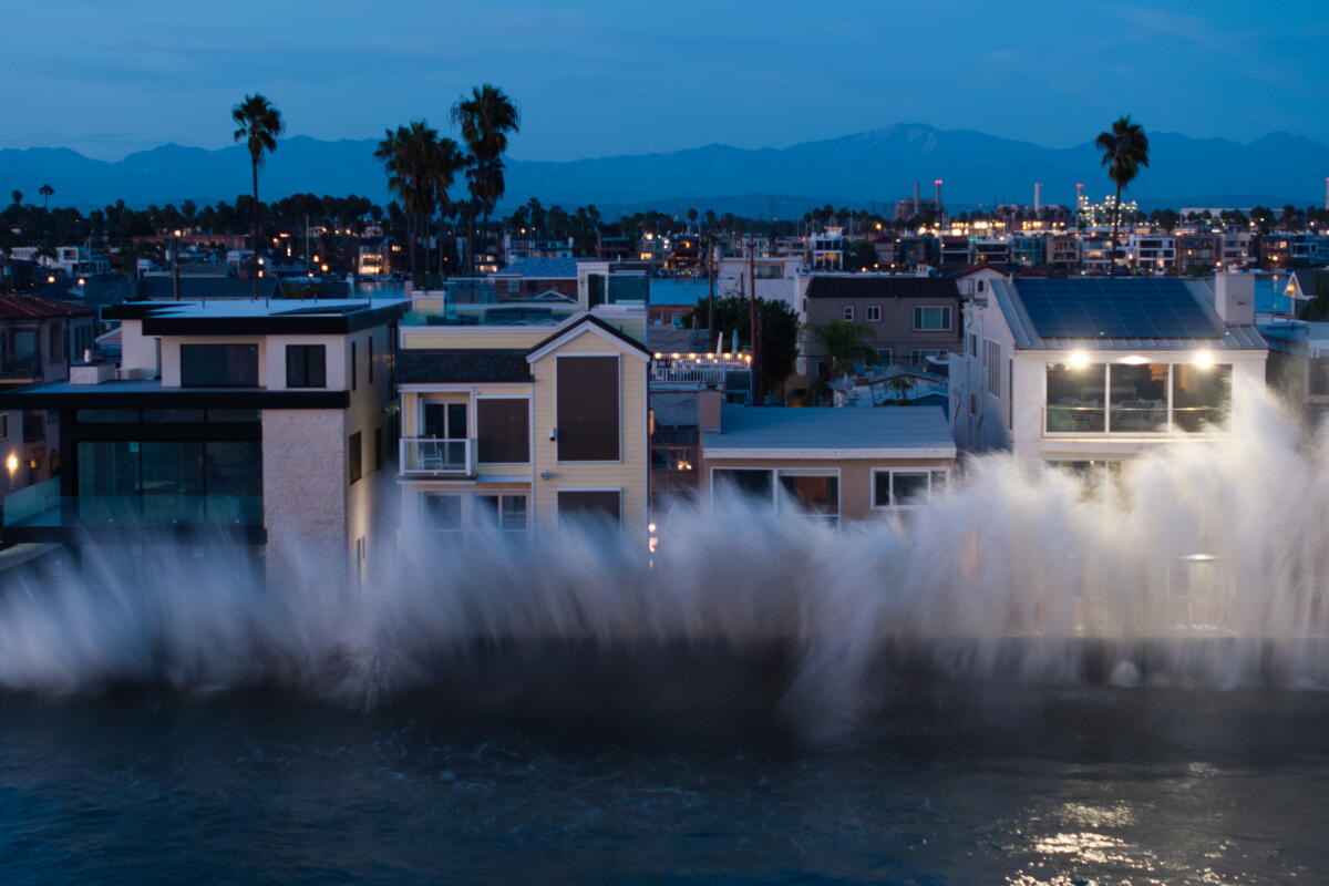 A large ocean wave hits a row of close-set, multi-story homes at nightfall.