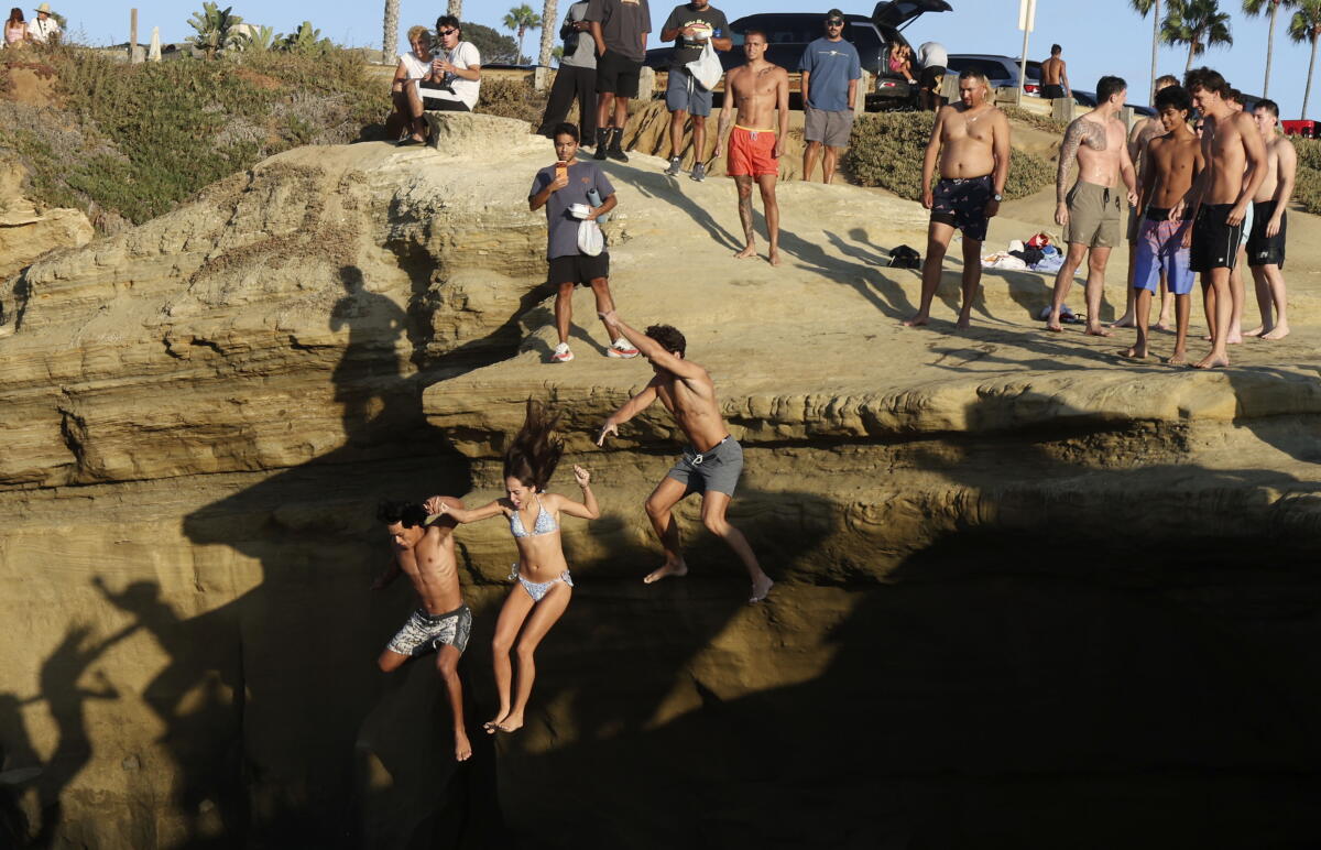 Three people in swimwear jumping from a cliff as spectators wait their turn