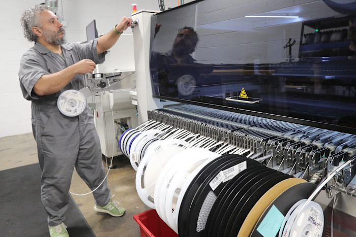 Dan Price prepares a pick and place machine, standing next to reels of electronic components in a factory setting.